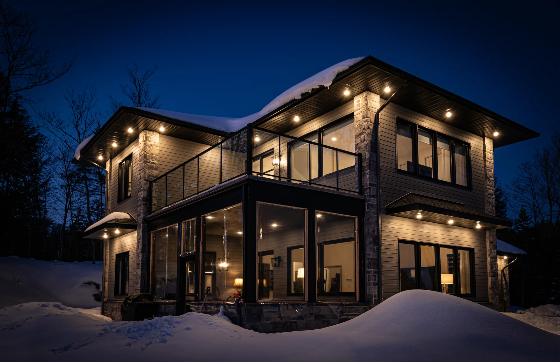 Résidence à Sainte-Adèle un soir d'hiver : toit enneigé, colonnes de pierre, balcon vitré et intérieur éclairé